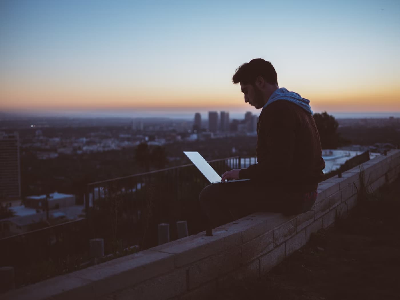 A man sitting on a ledge using his laptop at sunset.
